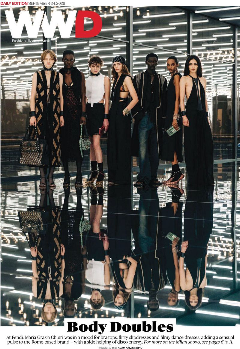 Seven fashion models standing in a row on a reflective glossy dark floor before a modern glass structure with linear overhead lighting.