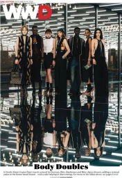 Seven fashion models standing in a row on a reflective glossy dark floor before a modern glass structure with linear overhead lighting.