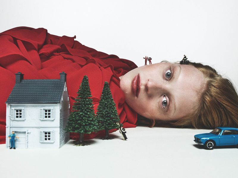 Surreal image with a giant woman lying next to miniature house, trees, car, and tiny people, all against a plain white background.