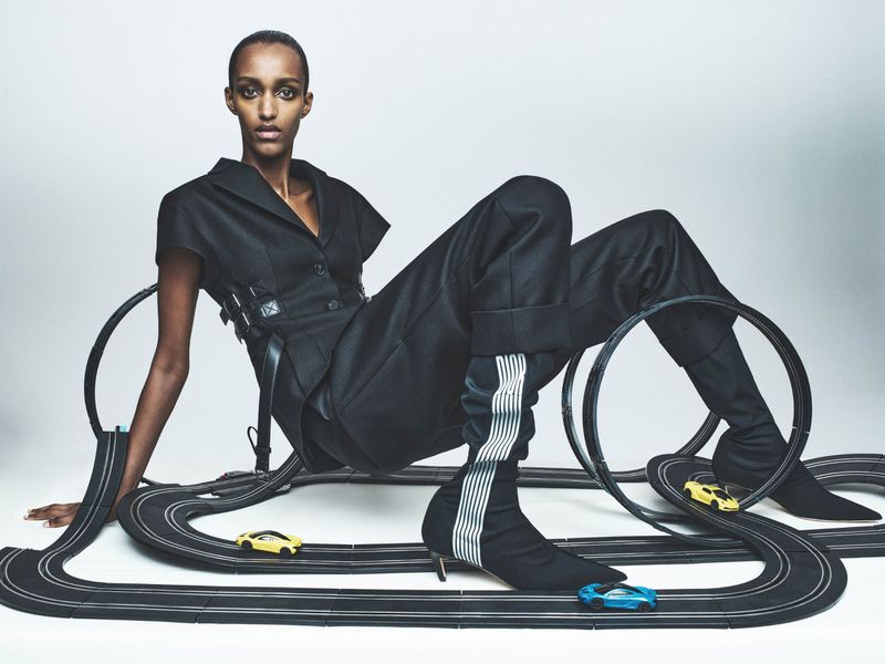 Fashion editorial shot of a black woman posing in a dark pantsuit next to a toy race track with toy cars in a light studio.
