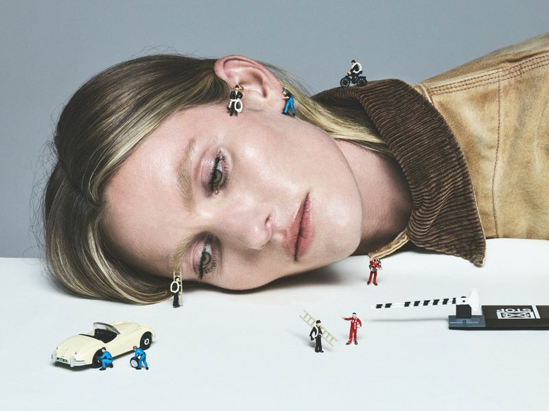 Conceptual beauty shot featuring a blonde woman's face at an angle with miniature figures and a toy car scattered around her.