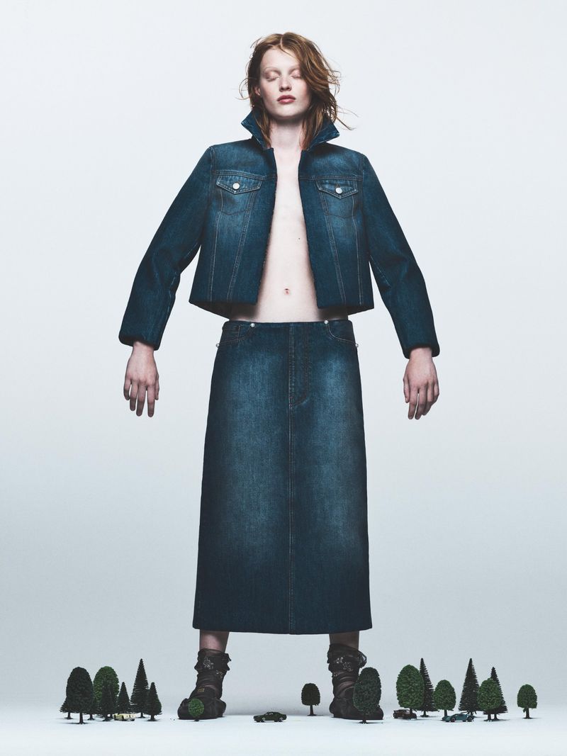 Fashion model in a denim jacket and skirt stands on a white background amid tiny trees, showcasing an editorial fashion aesthetic.