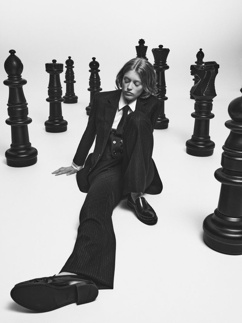 An adult in a pinstripe suit sitting on the floor surrounded by large black chess pieces against a plain white background in a studio.