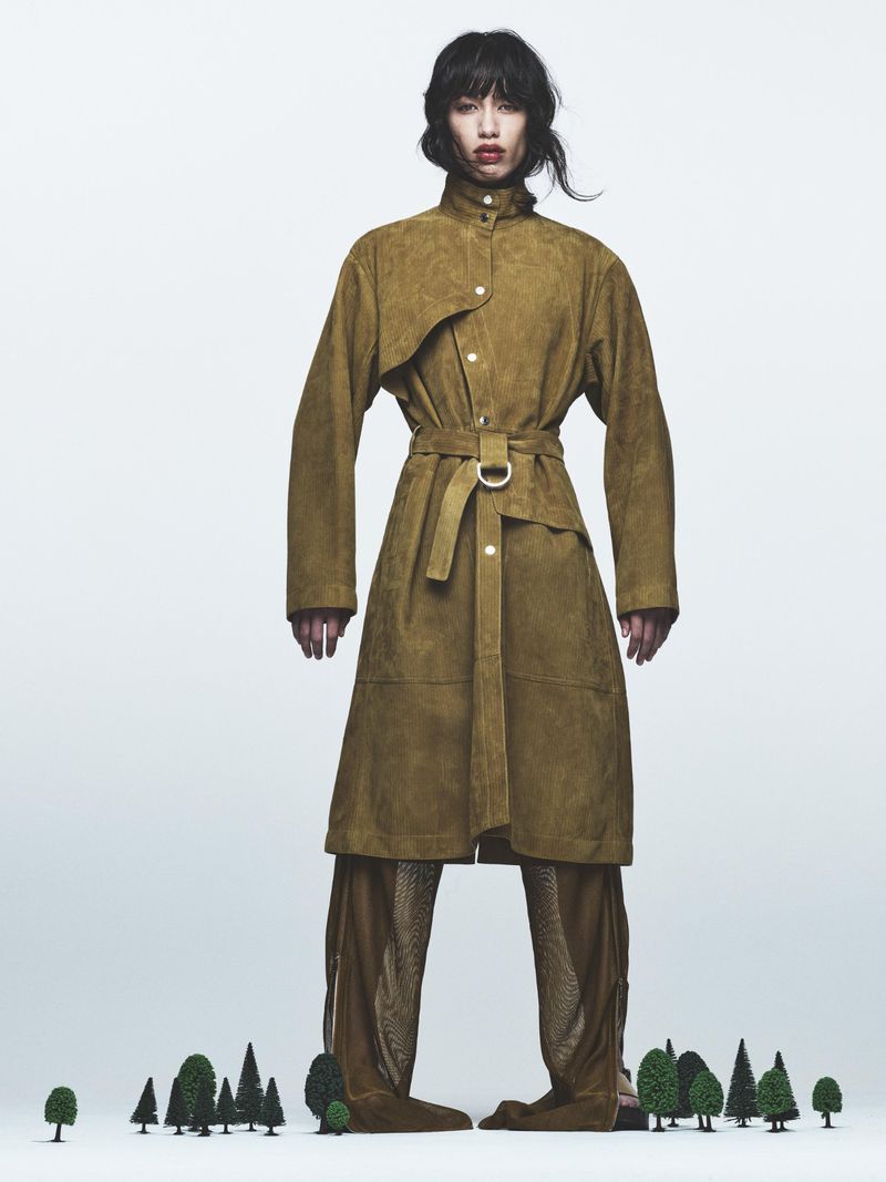 Fashion shot of a slender woman in a belted tan coat and textured brown pants standing on miniature green trees in a white studio.