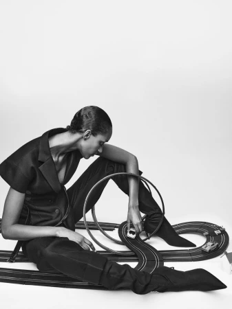 Woman in a dark suit poses next to a toy race track with small toy cars in a studio with soft lighting, shot in black and white.