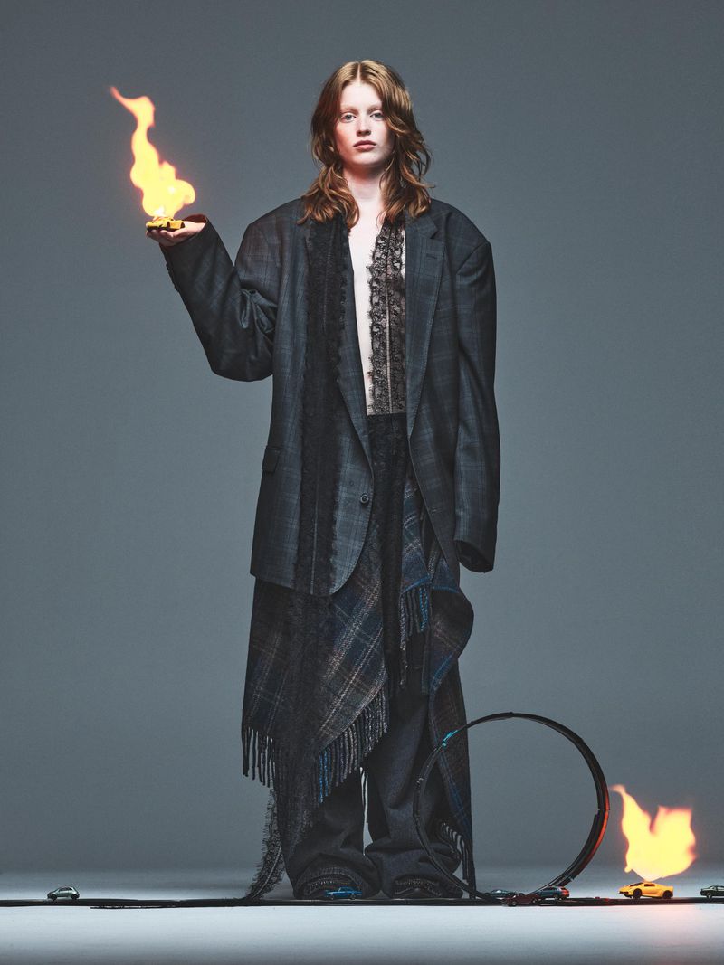 A fashion shot features a red-haired woman in an oversized blazer holding a small flame, with a burning loop in the foreground on gray.