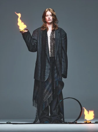A fashion shot features a red-haired woman in an oversized blazer holding a small flame, with a burning loop in the foreground on gray.
