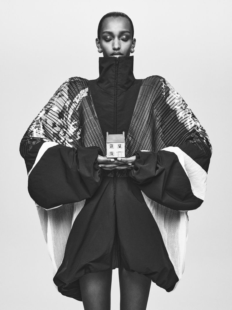 Fashion shot featuring an adult woman in an oversized jacket holding a miniature house. The photo is in black and white with studio lighting.
