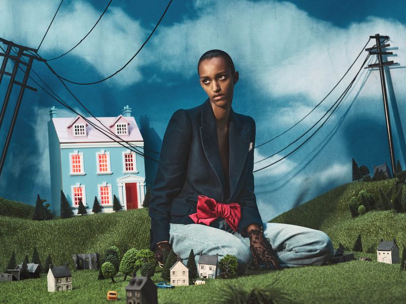 Fashion shot of a Black female model sitting in front of a miniature town and a blue house with a blue sky background.