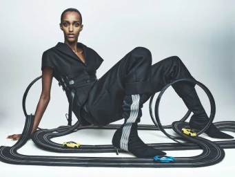 Model in black tailored jacket and trousers seated among toy race car tracks with miniature cars against a grey studio backdrop