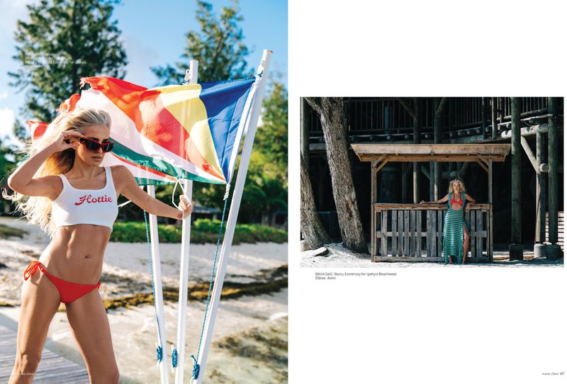Blonde woman in white crop top and red bikini bottoms posing by colourful flags on a sandy beach