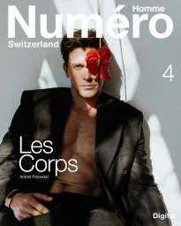 Antoni Porowski posing shirtless under a black jacket with a red flower covering one eye against a grey wall.