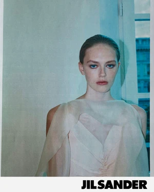 Model wearing a sheer light-colored sleeveless top against a pale interior background with blue lighting accents.