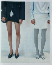 Two models standing side by side wearing skirts, tights, and pointed-toe heels against a plain white studio background.