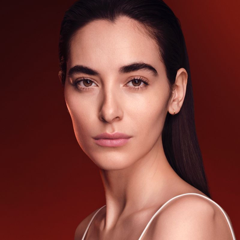 Close-up beauty portrait of a woman with slicked-back dark hair and subtle makeup against a warm dark red background.