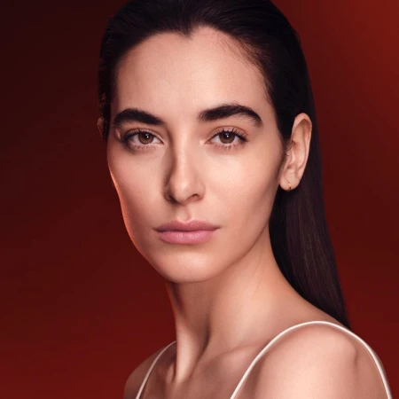 Close-up beauty portrait of a woman with slicked-back dark hair and subtle makeup against a warm dark red background.