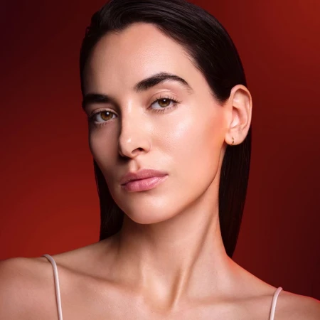 Close-up beauty portrait of a woman with slicked-back dark hair and glowing skin against a warm red gradient background