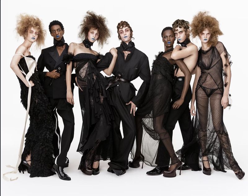 Group of models in black avant-garde fashion designs with voluminous hair and blue lipstick standing against a white backdrop.