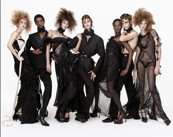 Group of models in black avant-garde fashion designs with voluminous hair and blue lipstick standing against a white backdrop.