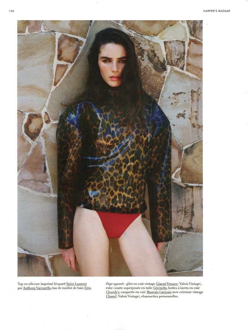 Fashion editorial shot of a slender woman in a leopard print top and red bikini bottoms posing against a stone wall indoors.