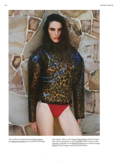 Fashion editorial shot of a slender woman in a leopard print top and red bikini bottoms posing against a stone wall indoors.