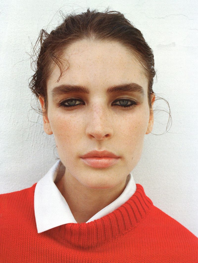 Close-up shot of a woman with light skin and dark eyes wearing a red sweater and a white collared shirt in front of a white wall.