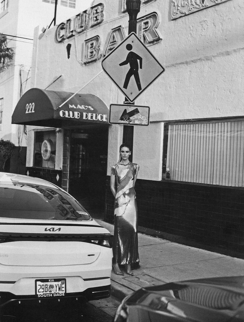 Fashion editorial shot of a model in a dress standing on a city sidewalk next to a white car and a crosswalk sign in black and white.