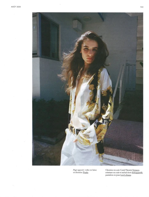 Fashion shot of a young adult woman with long brunette hair wearing a patterned white shirt and white pants standing outdoors.