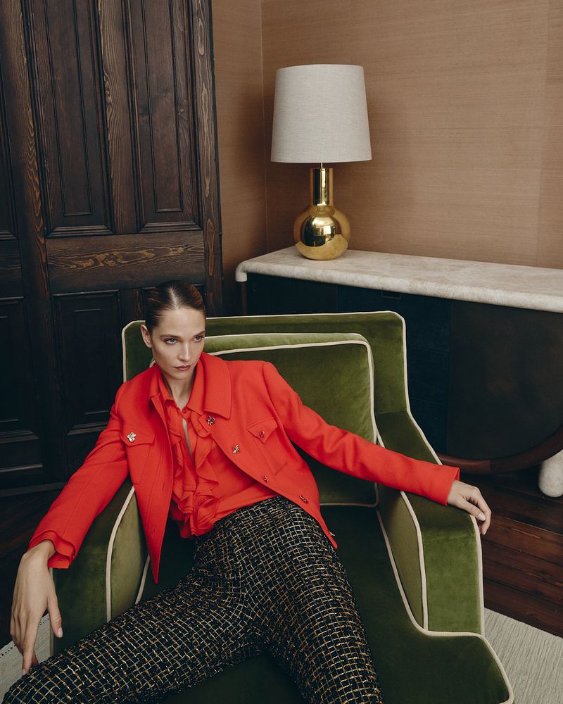 Model wearing a vibrant red jacket and tweed trousers seated in a green velvet armchair inside an upscale interior setting.