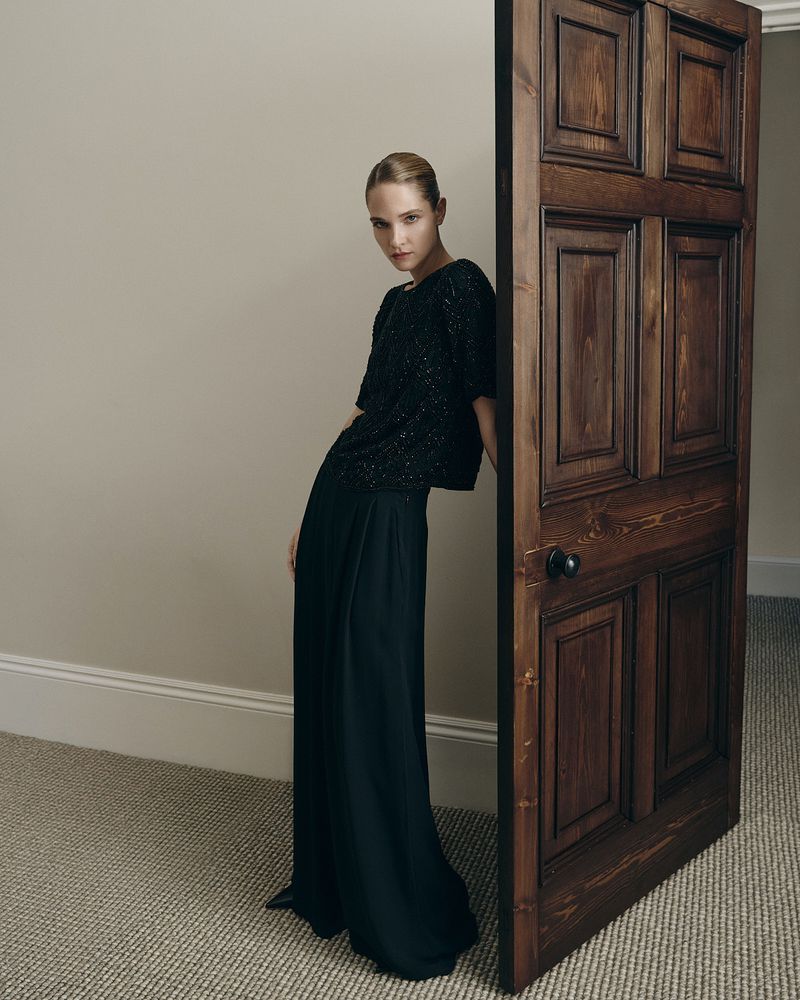 Model leaning against a dark wooden door wearing a black textured top and wide trousers in a room with a carpet