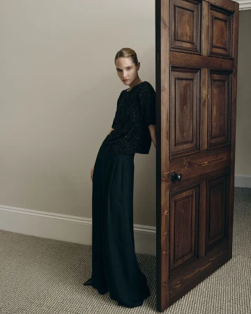 Model leaning against a dark wooden door wearing a black textured top and wide trousers in a room with a carpet