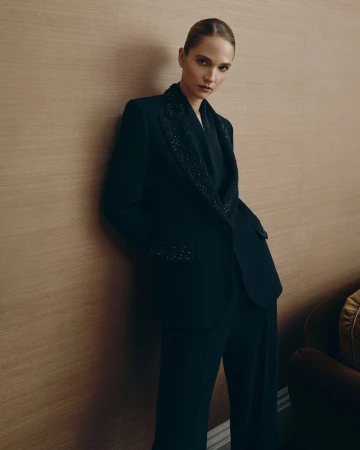 Model Barbara Kovacs leaning against a textured wall wearing a dark embellished trouser suit for Louise Kennedy AW26