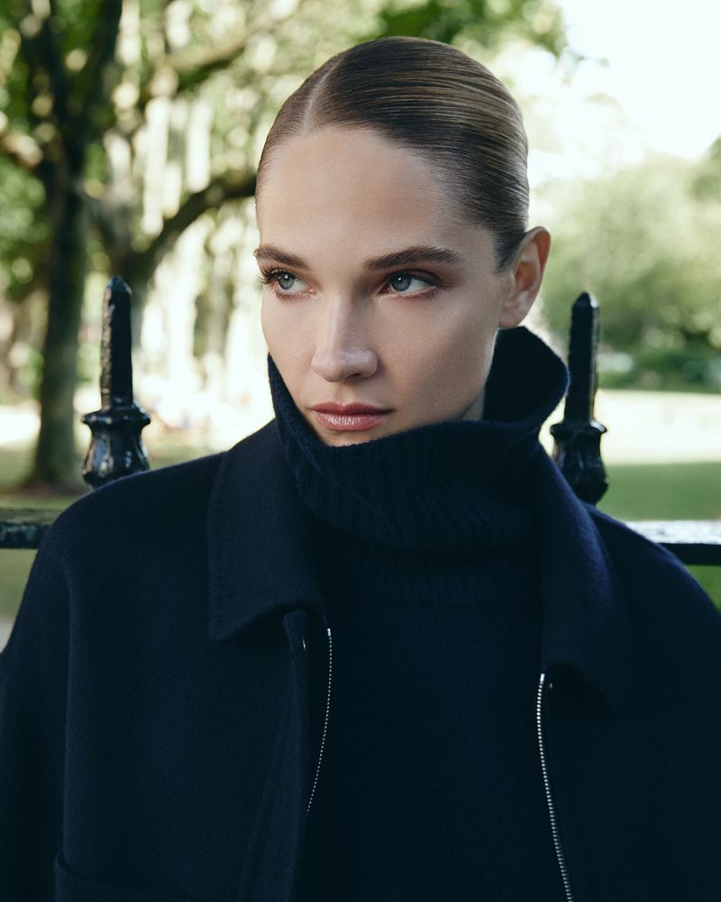 Model with slicked-back hair wearing a dark high-collar coat outdoors with a blurred park background.