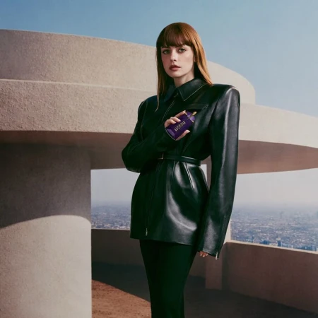 Woman in a structured black leather jacket holding a purple perfume bottle on a concrete rooftop overlooking a city skyline