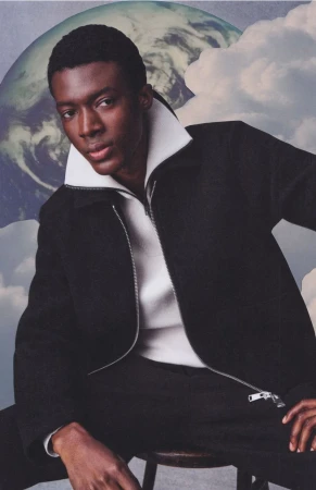 Black male model wearing a dark zipped jacket over a white collar against a surreal cloud and planet backdrop