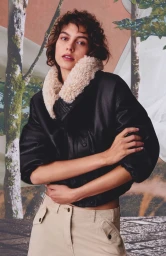 Model wearing a black leather jacket with a fluffy collar and beige trousers against a forest backdrop
