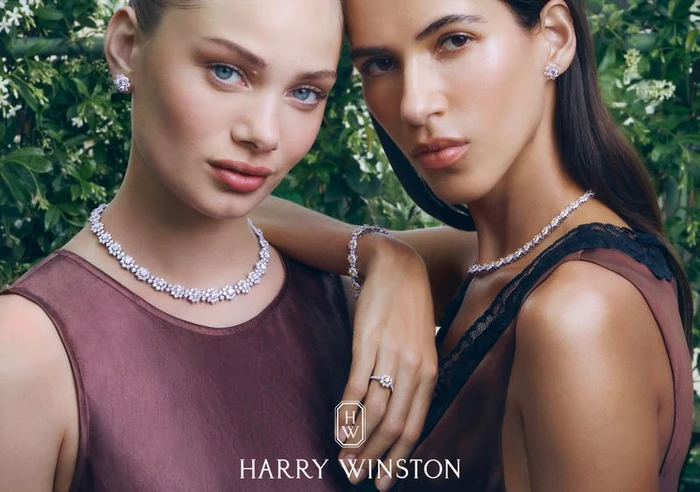 Two female models wearing diamond necklaces and earrings against a lush green garden background for Harry Winston.