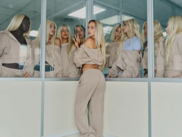 Bad Gyal wearing matching beige fleece hoodie and sweatpants posing in an office setting surrounded by glass partitions.