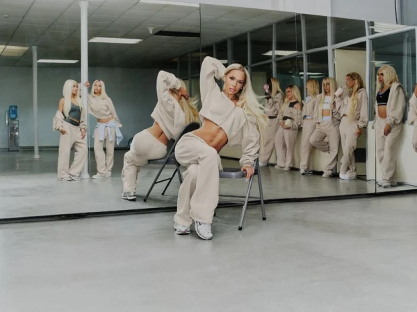 Bad Gyal wearing beige Nike fleece hoodie and sweatpants posing on a chair in a mirrored dance studio