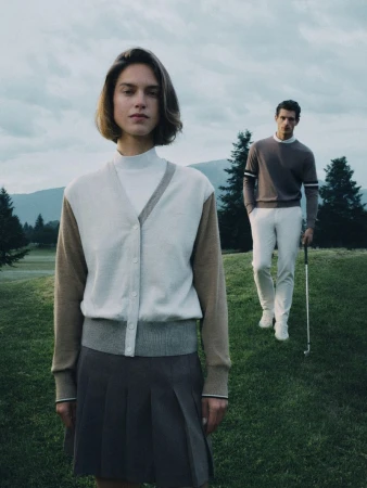 Woman in foreground wearing a cardigan and pleated skirt with a man holding a golf club on a grassy course behind her for HUGO Golf.