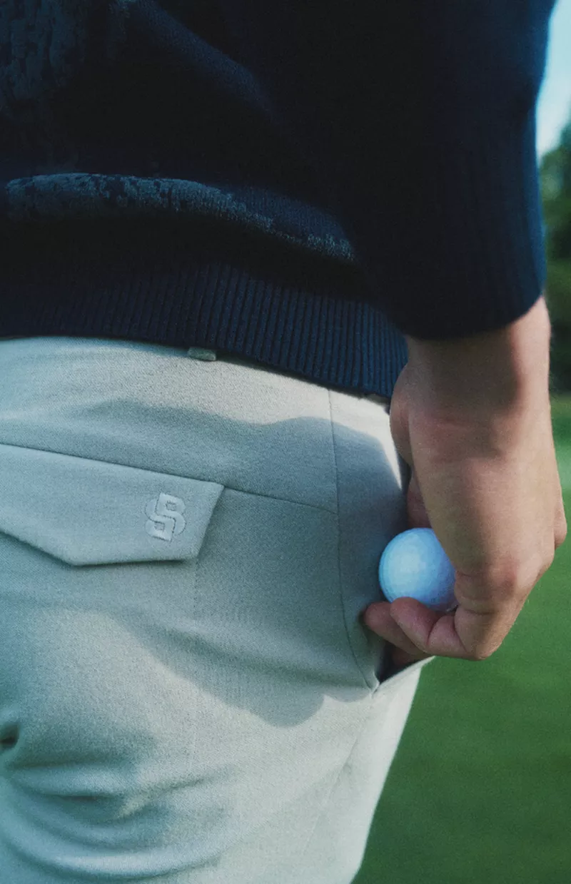 Close-up of a person holding a golf ball near their pocket while wearing light trousers and a dark sweater on a course