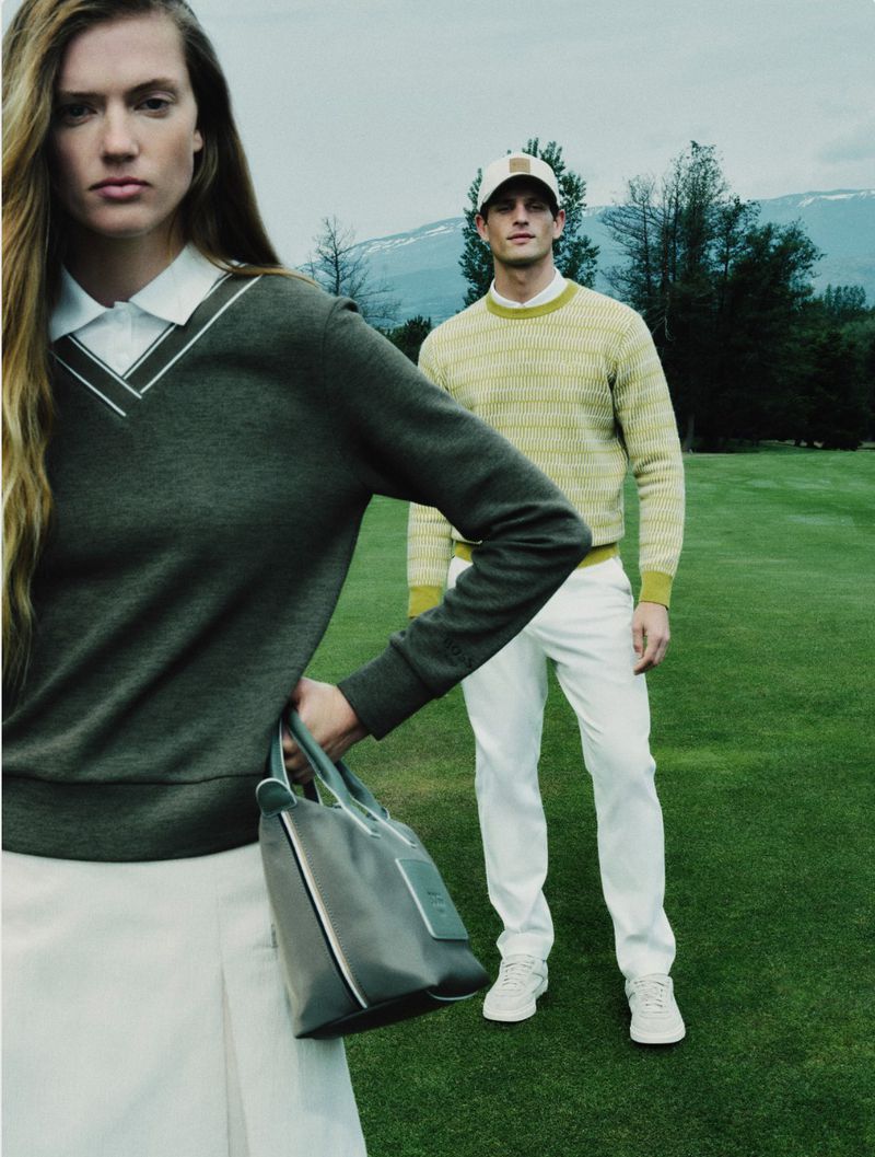 Model in dark green v-neck sweater and white skirt holding a grey handbag on a golf course with a male model in the background