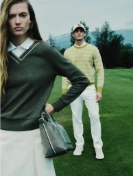 Model in dark green v-neck sweater and white skirt holding a grey handbag on a golf course with a male model in the background