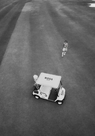 High-angle monochrome view of a golfer walking on a course near a branded golf cart.