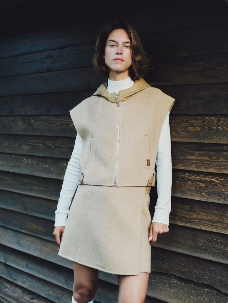 Model standing against a dark wooden wall wearing a beige hooded vest and matching skirt over a white turtleneck.