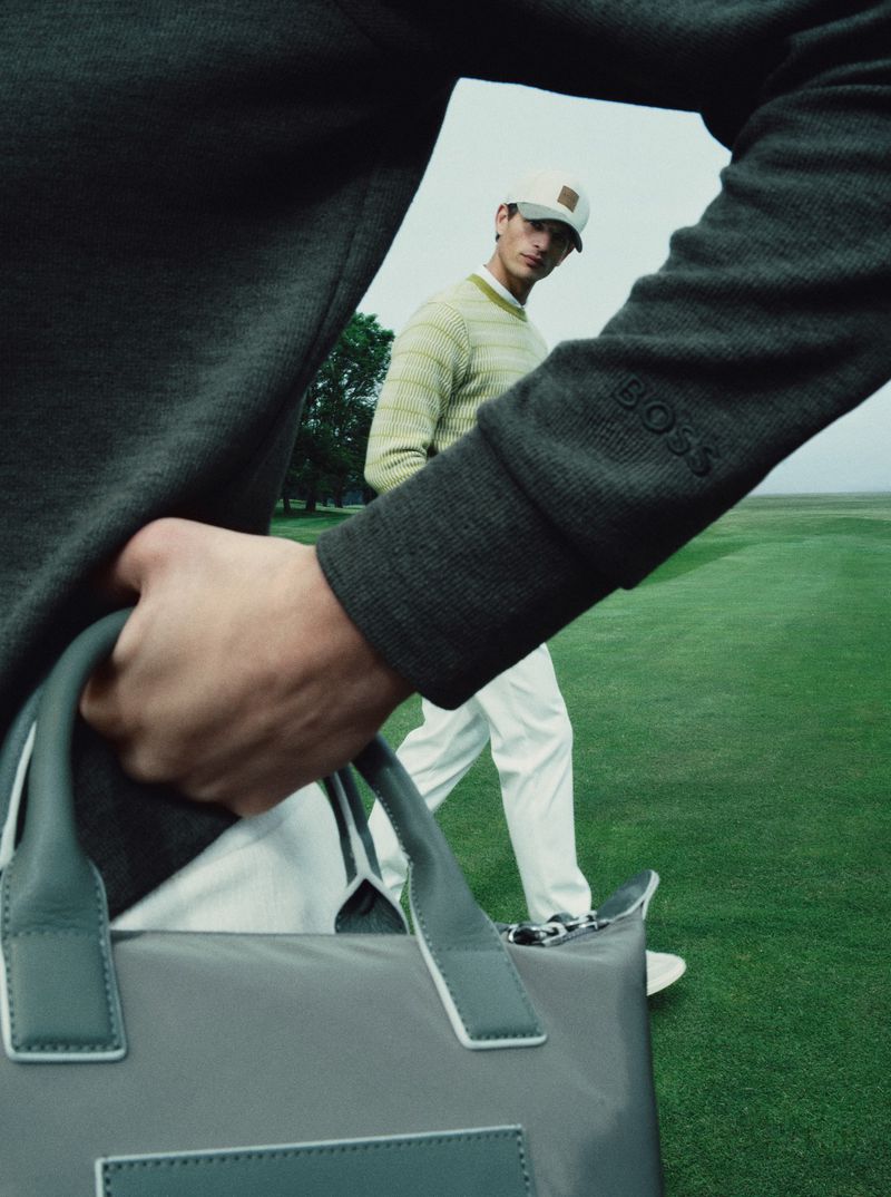 Man wearing a green long-sleeve sweater and white cap walking on a golf course while holding a grey handbag.