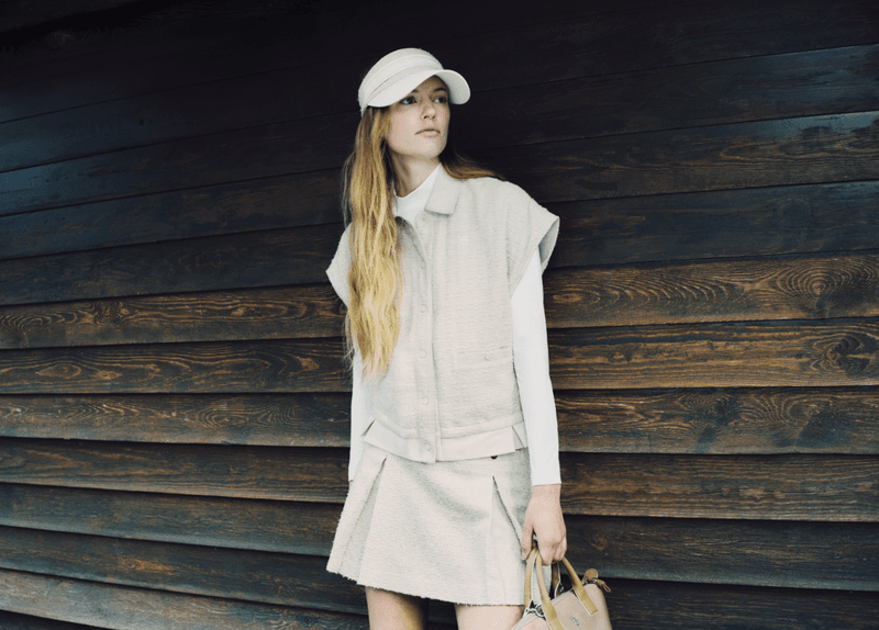 Model wearing a white visor, sleeveless vest, and skirt leaning against dark wood siding