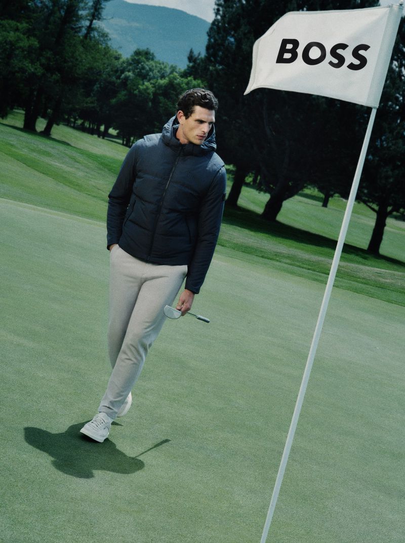 Man wearing dark hooded puffer jacket and light trousers walking on a green golf course near a white flag