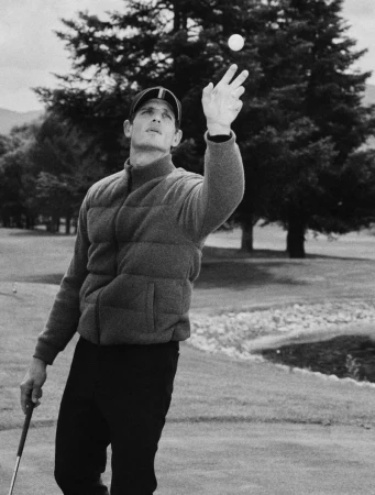 Man wearing a cap and puffer jacket tossing a golf ball in the air on an outdoor course with trees in the background