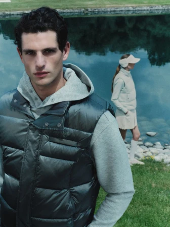 Close-up of a man in a quilted puffer vest and grey hoodie outdoors near a body of water with a woman standing in background.